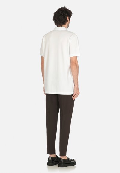 Uomo che indossa una polo bianca a maniche corte, pantaloni scuri su misura e scarpe di pelle nere, in piedi di spalle su uno sfondo bianco.