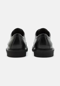 Par de zapatos de vestir de cuero negro mostrados desde atrás sobre un fondo claro y liso.