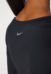 Crna atletska majica s širokim izrezom. Ima svijetlosivi Nike logo smješten na gornjem dijelu leđa, glatku teksturu i prilagođen dizajn.