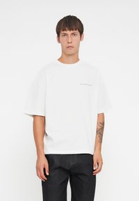 Witte katoenen t-shirt met korte mouwen en een ronde halslijn. Bevat subtiele grijze tekst "VERSCHWEND GEEN CULTUUR" op de linkerborst.