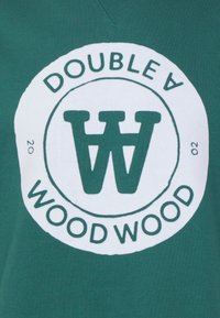 Zelený oděv s kruhovým logem, nad nímž je nápis „DOUBLE A“ a pod ním „WOOD WOOD“ se stylizovaným písmenem „W“. Látka vypadá hladká s jemným leskem.