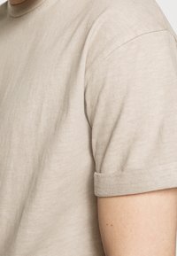 Světle béžové tričko s krátkým rukávem a kulatým výstřihem. Měkká látka s hladkou texturou a detailním lemováním rukávů.