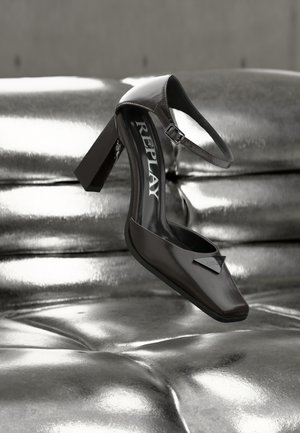 Fekete bőr magassarkú cipő bokapánttal és "REPLAY" márkajelzéssel, fényes, fémes ezüst színű, rikító párnázott felületen kiállítva.