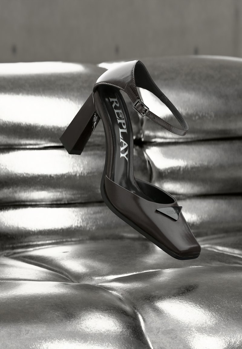 Μαύρο δερμάτινο παπούτσι με ψηλό τακούνι και λουράκι στον αστράγαλο με την επιγραφή "REPLAY", τοποθετημένο πάνω σε μια γυαλιστερή μεταλλική ασημί επιφάνεια με πλούσια επένδυση.