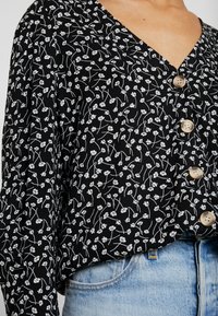 Blusa nera con stampa floreale, dotata di scollatura a V, maniche lunghe e bottoni in legno, realizzata in tessuto leggero con una texture morbida.