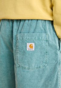 Türkise Corduroy-Shorts mit vertikalen Rippungen, ausgestattet mit einer Gesäßtasche und einem weißen Etikett mit einem orangefarbenen Logo. Gelbes Oberteil teilweise sichtbar.