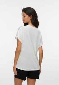 Mujer con cabello largo y oscuro que lleva un top blanco de manga corta con detalle de encaje en el hombro y pantalones cortos negros, de espaldas.