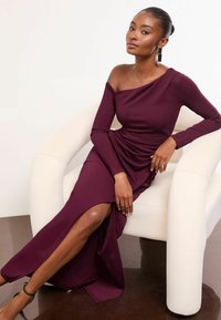 Burgundska haljina s otkrivenim ramenima, dugim rukavima, drapiranim materijalom, izrezom do butina i glatkom teksturom, uparen s bijelom stolicom i sjajnim podom.