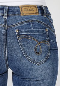 Gros plan sur la poche arrière d'un jean en denim bleu avec des coutures décoratives, des rivets et une étiquette de marque en cuir marron portant la mention « Koroshi Denim Collection ».