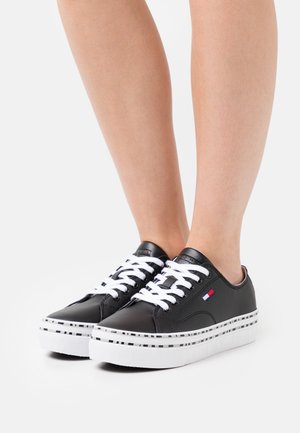 Nohy obuté v černých nízkých teniskách s bílými tkaničkami a vzorovanou bílou podrážkou, na boku s malou vlajkovou nálepkou v červené, bílé a modré barvě.