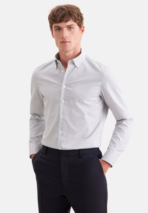Junger Mann steht mit den Händen in den Taschen, trägt ein weiß-grau kariertes Hemd mit Knöpfen und dunkle Hosen vor weißem Hintergrund.