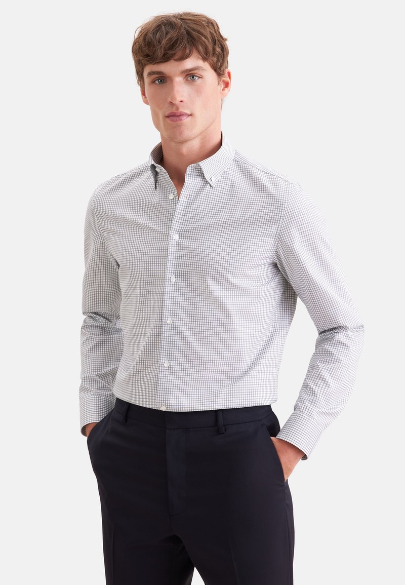 Jeune homme debout les mains dans les poches, portant une chemise blanche et grise à carreaux boutonnée et un pantalon sombre sur fond blanc.