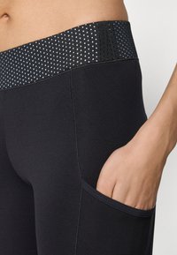 Czarne legginsy sportowe z teksturowanym pasem w białe kropki, boczną kieszenią i gładkim materiałem, zaprojektowane dla komfortu i swobody ruchów.