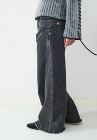 Szerokie czarne dżinsy z postrzępionym dołem. Posiadają kieszenie boczne oraz unikalny skośny design kieszeni z przodu. Widoczna faktura materiału.