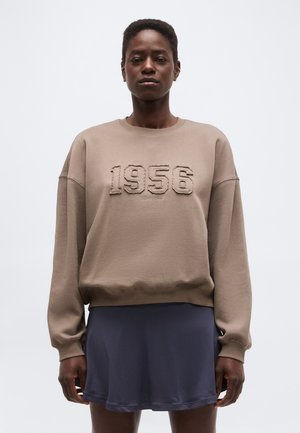 Άτομο που φοράει φούτερ χρώματος ταουπέ με το κείμενο "1956" και σκούρα ναυτική φούστα, στέκεται μπροστά σε απλό ανοιχτό γκρι φόντο.