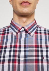 Homme portant une chemise à carreaux rouge, blanche, marine et bleue, boutonnée avec un col boutonné et un petit logo Tommy Hilfiger sur la poitrine.