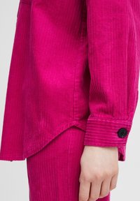 Fuchsia Kordhemd mit strukturiertem Finish, langärmlig, mit spitzem Saum und einem schwarzen Knopf an den Manschetten.