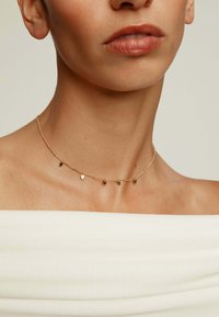 Collarín de oro con pequeños acentos en forma de corazón, cadena delicada y un acabado suave, que se lleva sobre una blusa blanca de hombros descubiertos.