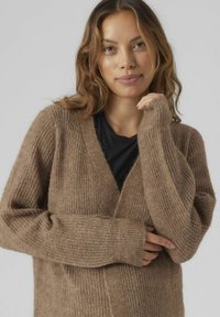 Cardigan marron en maille texturée, avec un col en V plongeant, des manches longues et des poignets côtelés, porté de manière décontractée sur une chemise noire.