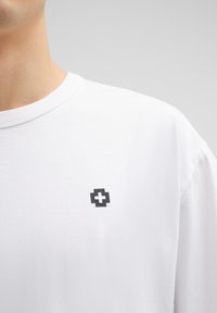 Witte katoenen t-shirt met een ronde hals en korte mouwen. Heeft een klein zwart logo op de linkerborst. Zachte textuur.