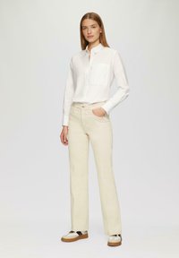Camicia bianca leggera con taschino frontale, abbinata a pantaloni a vita alta color crema e sneakers beige con dettagli neri.