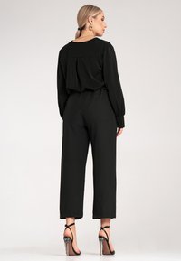 Czarny kombinezon o luźnym kroju, z długimi rękawami i plisowanym detalem na plecach. Zestawiony z przezroczystymi sandałami na wysokim obcasie.