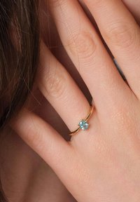 Goldring mit einem hellblauen ovalen Edelstein, mit einem glatten Band und vier Krappen, die den Stein halten, vor einem Handhintergrund.