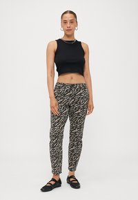 Top corto nero, pantaloni stampati beige e neri, e scarpe nere. La modella è in piedi su uno sfondo bianco. Orecchini a cerchio in argento.