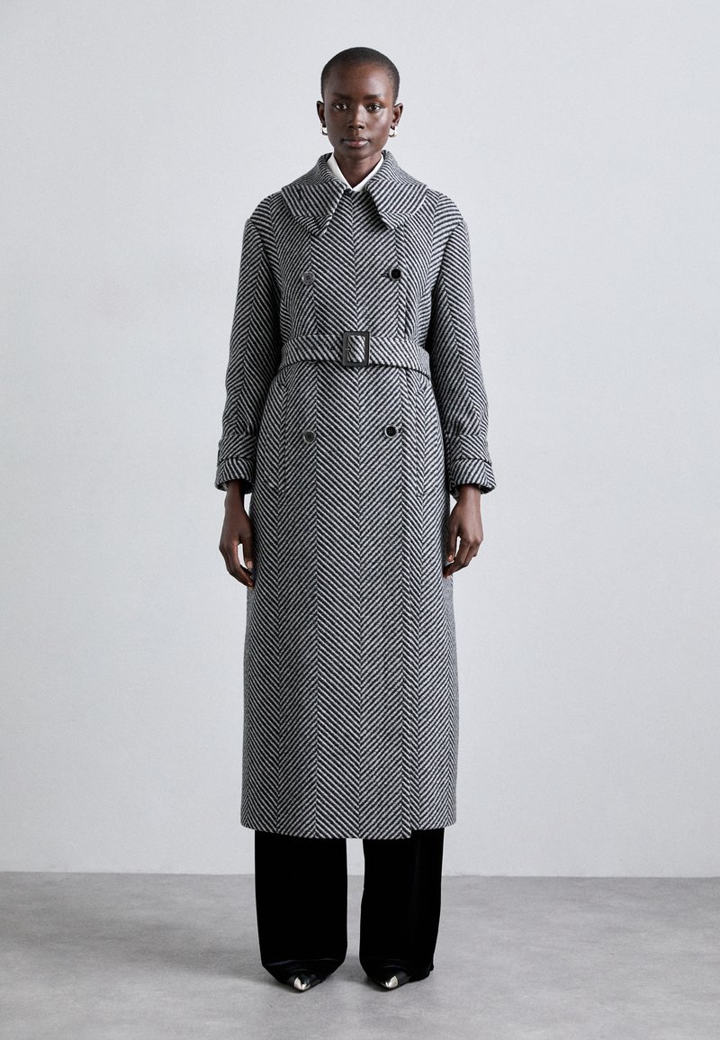 Schwarz-weißer Fischgrät-Trenchcoat mit breitem Kragen, doppelreiheigen Knöpfen und einem Gürtel in der Taille, kombiniert mit schwarzen Hosen und Schuhen.