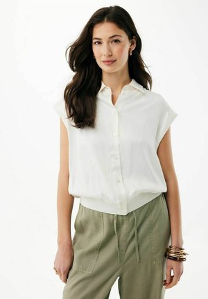 Donna con capelli lunghi e scuri che indossa una camicia bianca senza maniche con bottoni e pantaloni verde oliva con coulisse, accessoriata con braccialetti e orecchini.