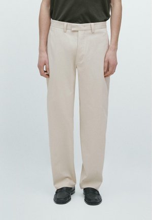 Mand iført beige bukser med lige ben, sorte sko og en mørk skjorte indeni bukserne, stående mod en ensfarvet baggrund.