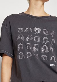 Mörkgrå T-shirt med vita konturteckningar av olika frisyrer och ordet "BOSS" längst ner till höger.