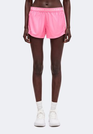 Személy rózsaszín Under Armour sport rövidnadrágot, fehér zoknit és fehér tornacipőt visel, egyszínű háttér előtt áll.