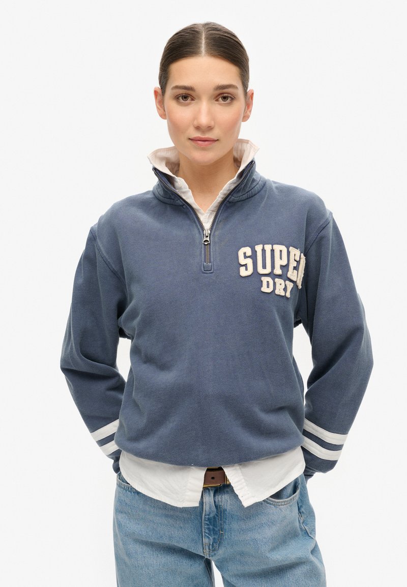 Veste à zip bleue avec l'inscription "SUPER DRY", accents blancs sur les manches et un tissu doux et texturé. Portée sur une chemise blanche à col.