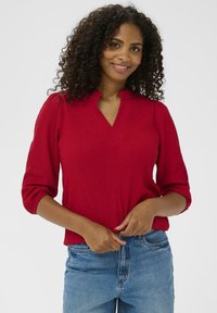 Rød ribbet strikbluse med henley-udskæring og trekvartærmer, parret med blå høj talje jeans. Glat tekstur og tætsiddende design.