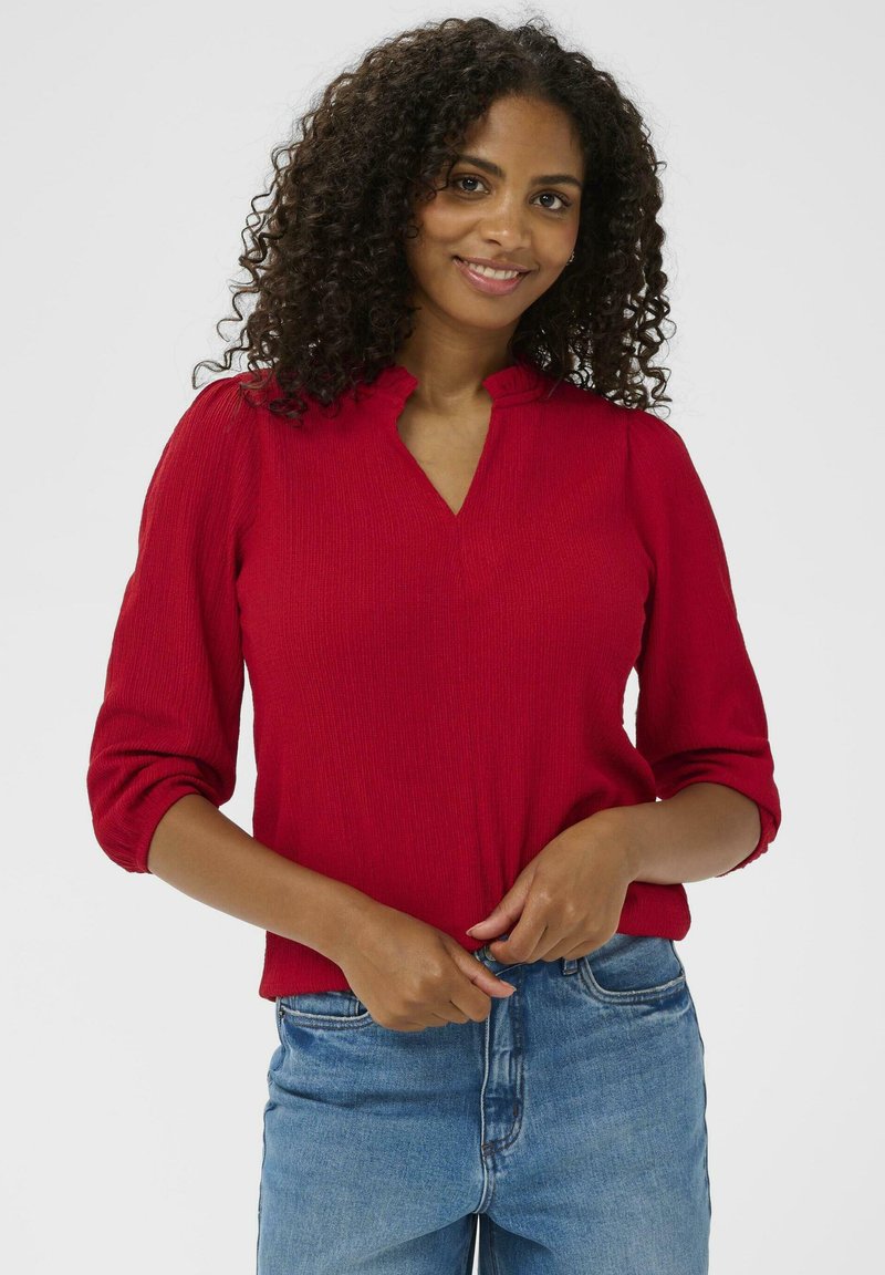 Rød ribbet strikbluse med henley-udskæring og trekvartærmer, parret med blå høj talje jeans. Glat tekstur og tætsiddende design.