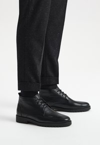 Czarne skórzane botki za kostkę o gładkiej fakturze, okrągłym nosku i sznurowanym przodzie, zestawione z ciemnymi spodniami w wzory.