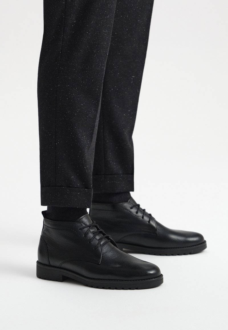 Czarne skórzane botki za kostkę o gładkiej fakturze, okrągłym nosku i sznurowanym przodzie, zestawione z ciemnymi spodniami w wzory.