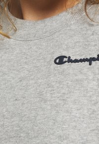 Sudadera de felpa gris con cuello redondo acanalado, que presenta un logotipo bordado en azul oscuro de "Champion" en el pecho. Tela suave y texturizada.