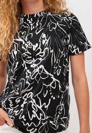 Femme aux cheveux bouclés portant un t-shirt noir avec un motif floral abstrait blanc et des manches courtes retroussées.