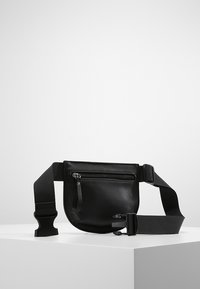 Musta nahkainen crossbody-laukku, jossa on vetoketjullinen etutasku, säädettävä kangashihna, pyöreä muoto ja sileä pinta.