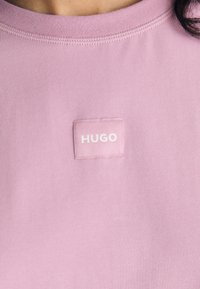 Lys rosa bomull T-skjorte med et kvadratisk merke med "HUGO"-tekst i hvitt. Rund hals med synlige sømdetaljer.