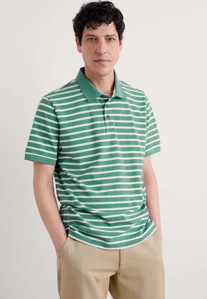 Grün gestreiftes Poloshirt mit weißen horizontalen Streifen, kurzen Ärmeln, einem Kragen und zwei Knöpfen am Hals; getragen mit beigen Hosen.