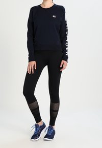 Felpa blu scuro con collo rotondo e branding bianco sulla manica, abbinata a leggings neri con dettagli in rete e scarpe da ginnastica blu.