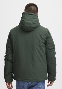 Mørkegrønn polstret jakke med hette, med en glatt tekstur, sidesømmer og en avrundet nederkant. Brukes over blå jeans.