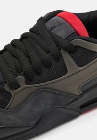 Sort sneakers med en tekstureret suede- og mesh-overdel, der har grå accenter og en rød detalje ved hælen. Inkluderer sorte snørebånd og en polstret sål.