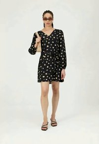 Γυναίκα φορώντας ένα μαύρο φορεμα με σχέδια και μακριά μανίκια, μαύρες σαγιονάρες με λουριά, γυαλιά ηλίου και κρατώντας μία μπεζ τσάντα ώμου.