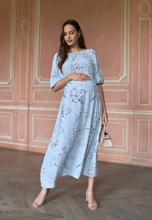 Femme enceinte portant une robe midi à fleurs bleu clair et des talons beiges, tenant un sac à main beige, debout dans une pièce élégante.