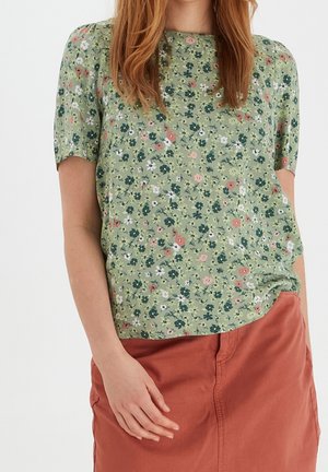 Mujer con blusa de flores de manga corta de color verde claro metida en una falda color óxido, de pie frente a un fondo blanco liso.