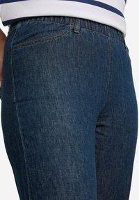 Mörkblå denimjeans med elastisk midja, som har en slät yta och klassisk femficksdesign. Sömmarna är synliga längs kanterna.